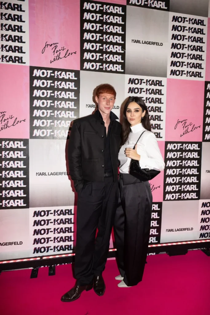 Two people pose on a pink carpet before a black, white and pink NOT-KARL checkerboard backdrop; one carries a black shoulder