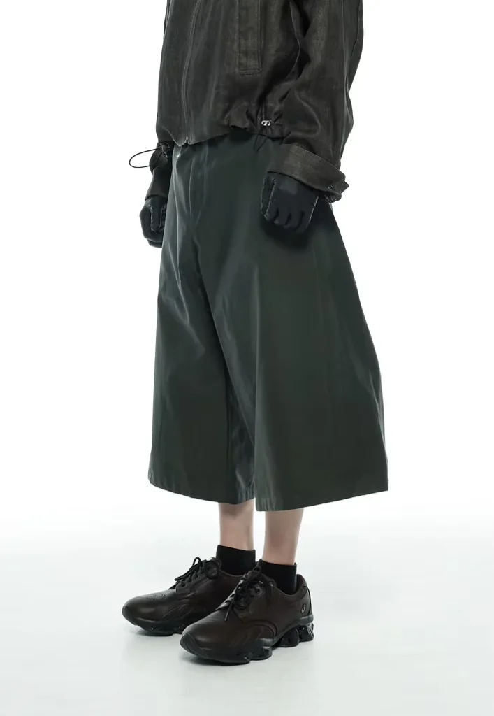 Cropped model wearing a black jacket, dark green knee-length shorts, gloves, socks and black sneakers against a white