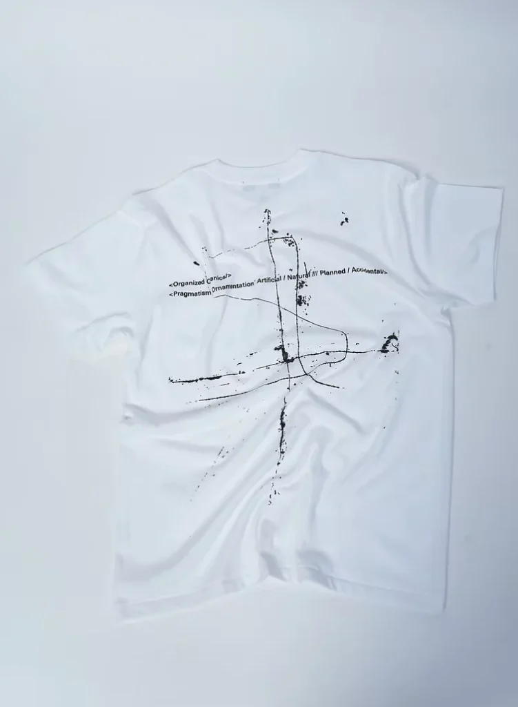 White Tee laid flat, featuring black abstract lines, splatters, and small printed text on a pale background