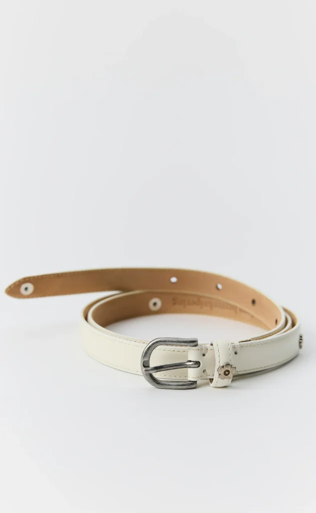 White leather belt coiled on a white surface, with a dark metal buckle and riveted strap details