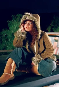 Remi Wolf in a fur-trimmed brown coat and hat sits on a vehicle, wearing blue jeans and tan boots