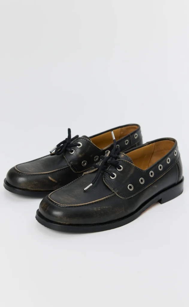 Pair of black leather boat shoes with laces, silver eyelets, tan lining and scuffed toes on a white background