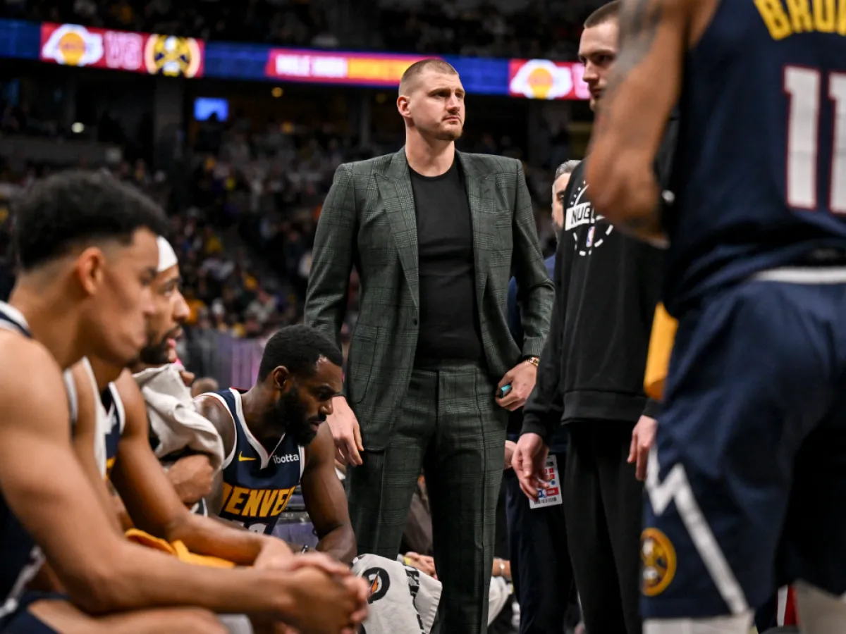 Nikola Jokić stands courtside in a gray plaid suit among seated Denver Nuggets players during a game