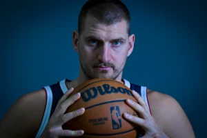 Nikola Jokic basketball player in a dark jersey holds an orange Wilson basketball against a blue background
