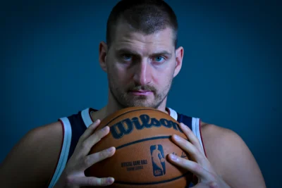 Nikola Jokic basketball player in a dark jersey holds an orange Wilson basketball against a blue background