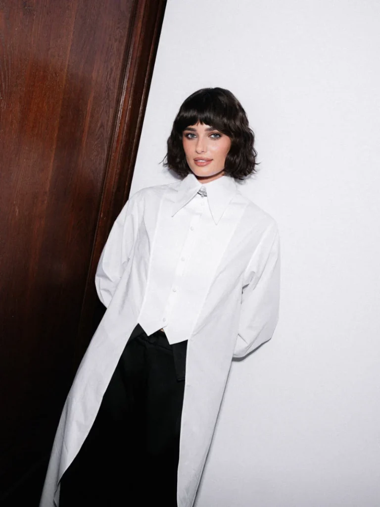 Woman with short dark hair poses in a white oversized shirt with pointed collar and black trousers beside a wooden door
