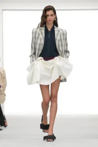 Model walks in a gray plaid blazer, navy knit top, white draped skirt, and black sandals on a runway