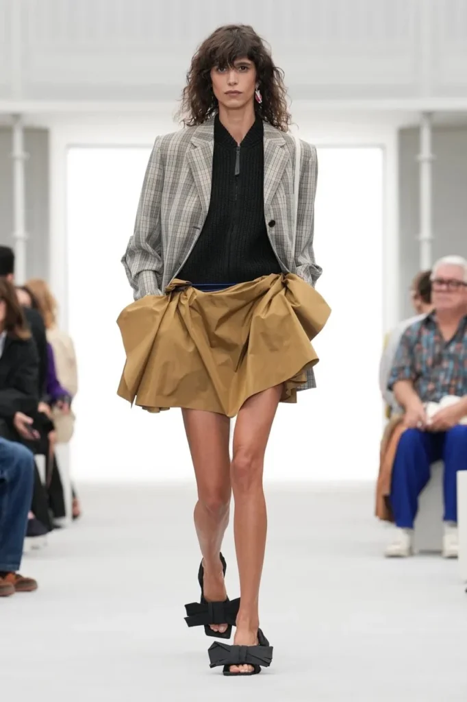 Model walks a runway in a gray plaid blazer, black sweater, draped tan skirt and black bow sandals