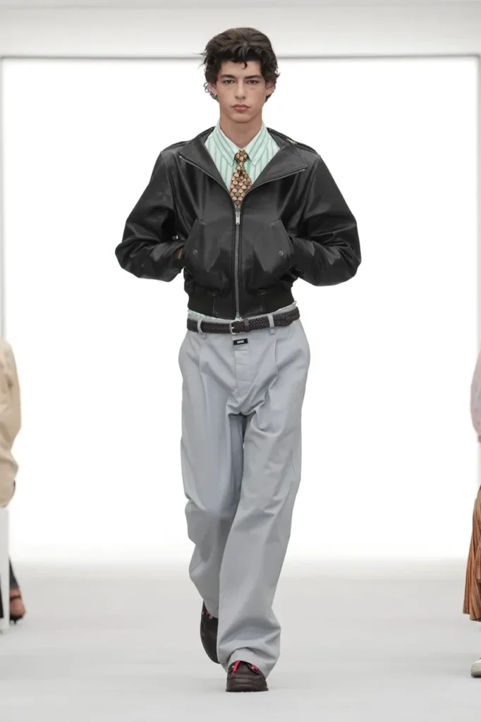 Model walking in a white runway space wearing a black leather jacket, striped shirt, patterned tie and loose gray trousers