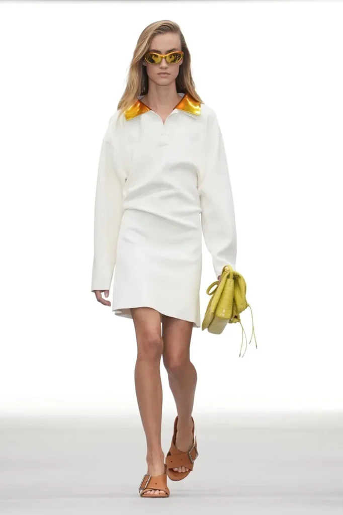 Model in white long-sleeved outfit with metallic orange collar, yellow sunglasses and handbag, walking against white
