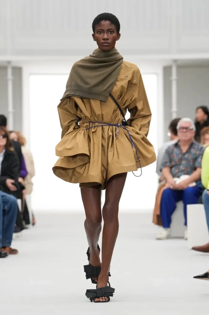 Model walks a runway in a tan cinched dress with layered skirt, olive scarf, blue cord belt and black strappy sandals