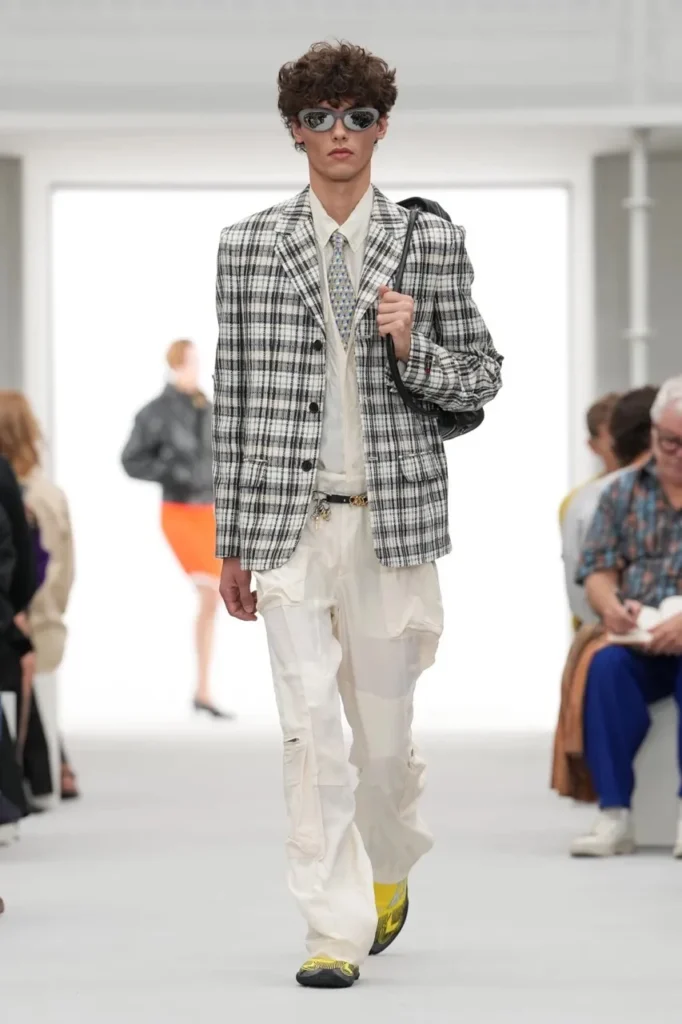 Model walks a runway in a black-and-white plaid blazer, white cargo pants, oval sunglasses and yellow-black shoes