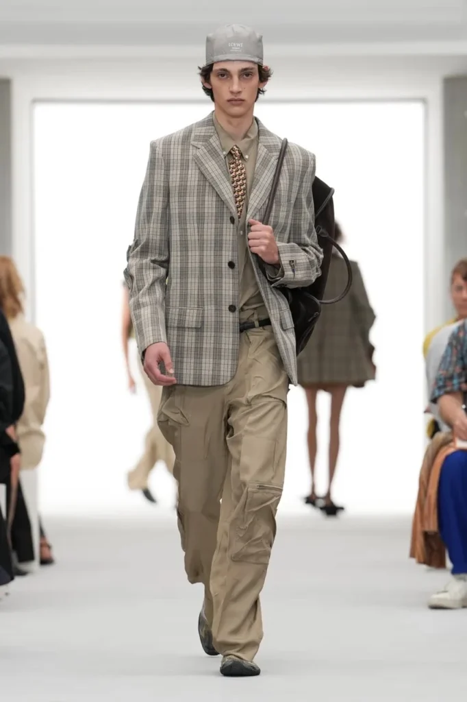 Model walks a runway in a gray plaid blazer, patterned tie, beige cargo trousers, cap, and black shoulder bag