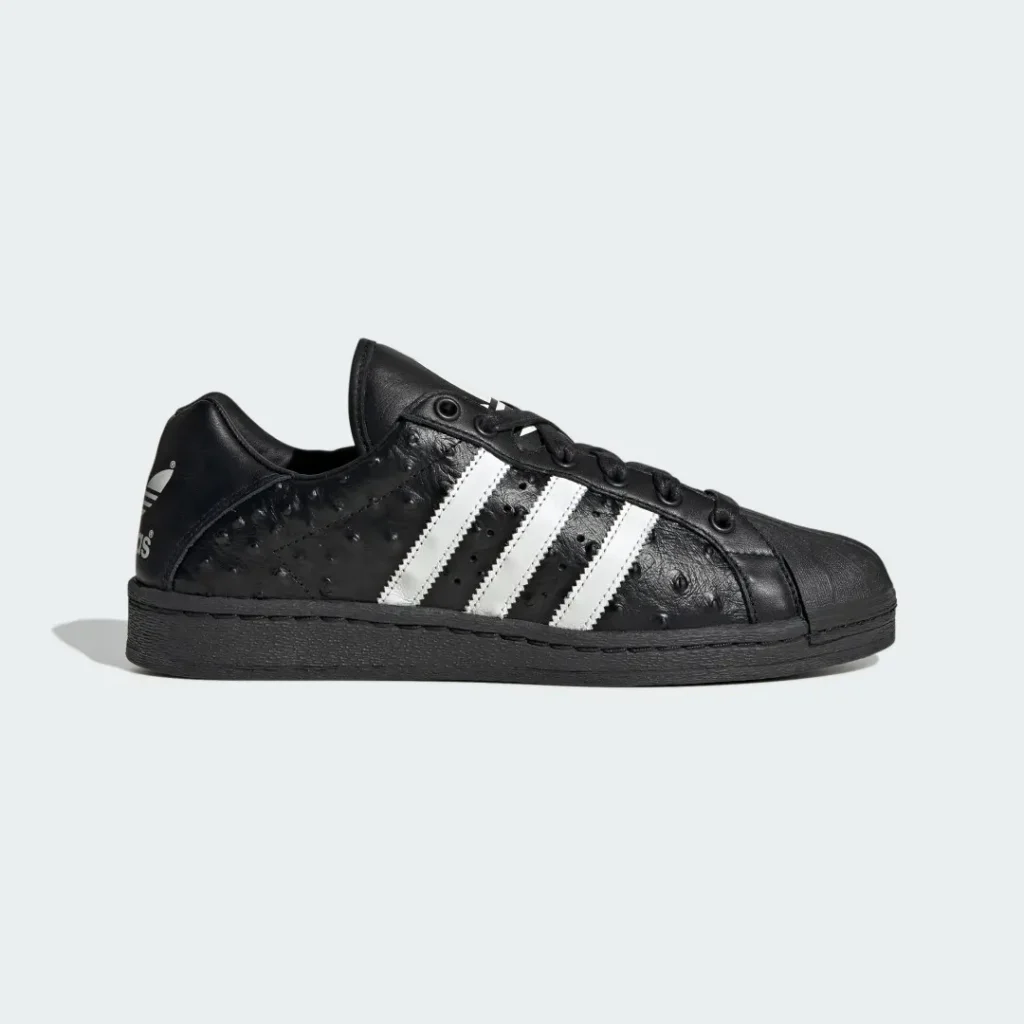 Black leather low-top sneaker with white three stripes, textured panels, black sole, and heel logo on a pale background