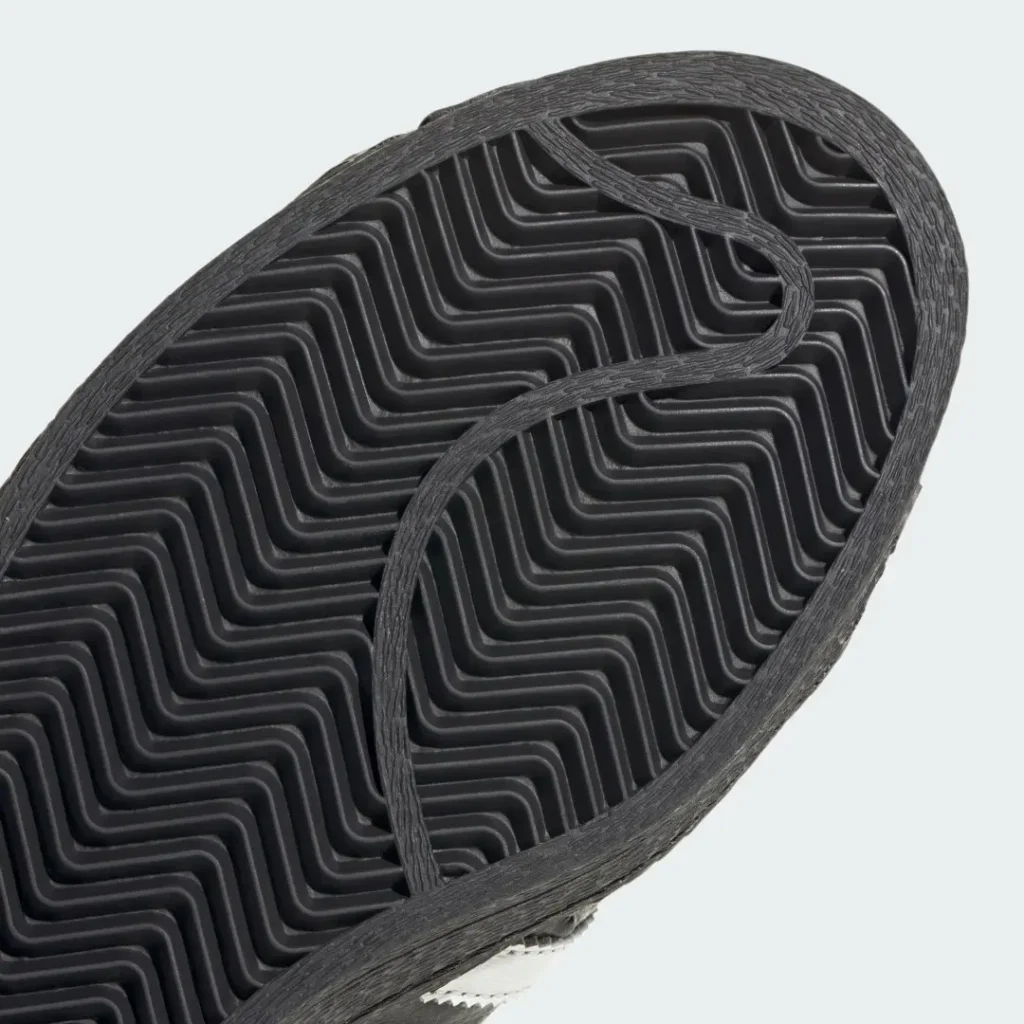 Close-up of a black rubber sneaker outsole with repeating zigzag tread and a textured raised border against a pale background