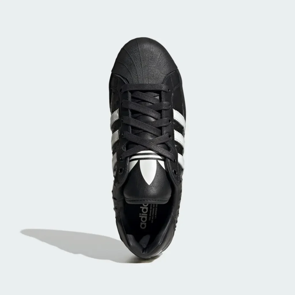 Top-down view of a black Adidas sneaker with white three stripes, laces, and a long shadow on a pale background