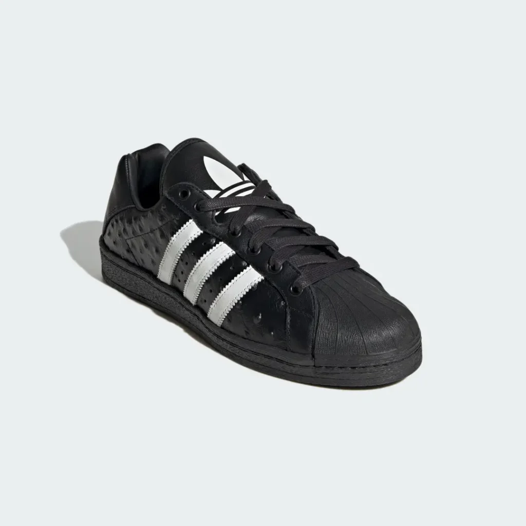 Black leather Adidas sneaker with white three stripes and shell toe, angled on a pale background