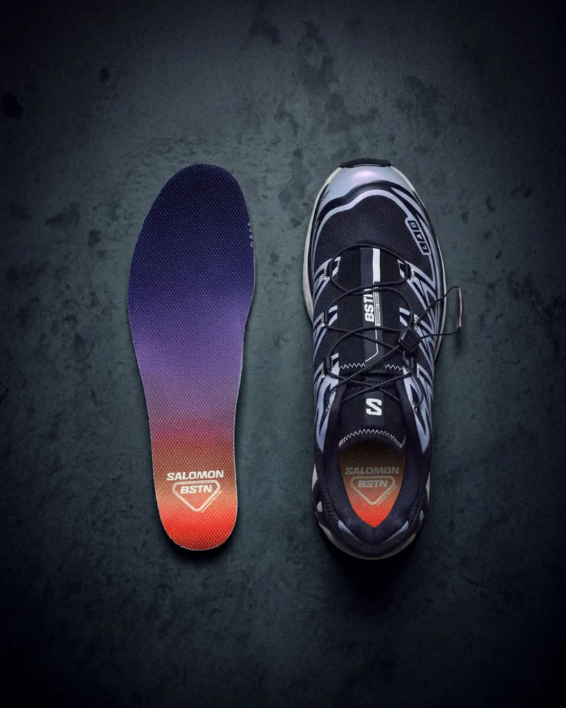 Top-down view of a black-and-white Salomon sneaker beside its purple-to-red gradient insole on a dark surface