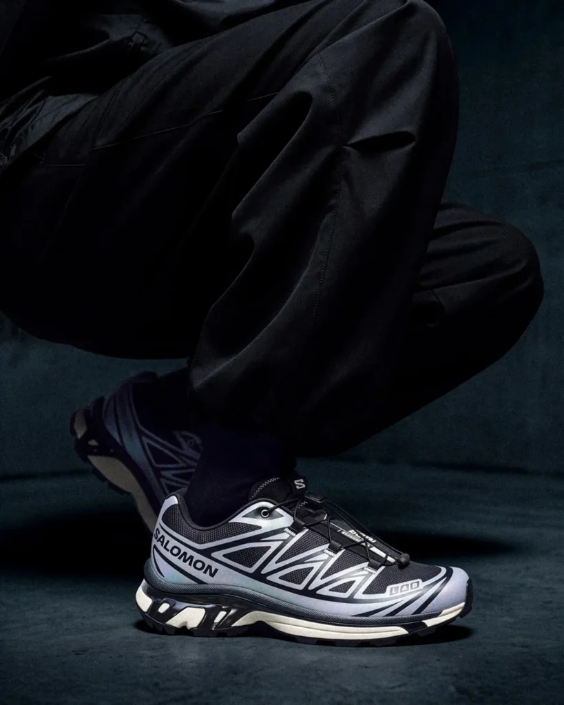 Close-up of a seated person in black clothing wearing gray-and-white Salomon XT-6 sneakers on a dark floor