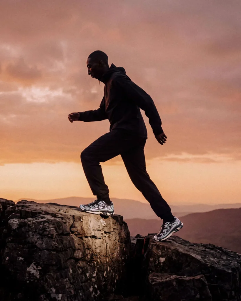 Person in black hooded jacket and pants strides across rocky cliffs wearing white and black trail shoes under a pink sky