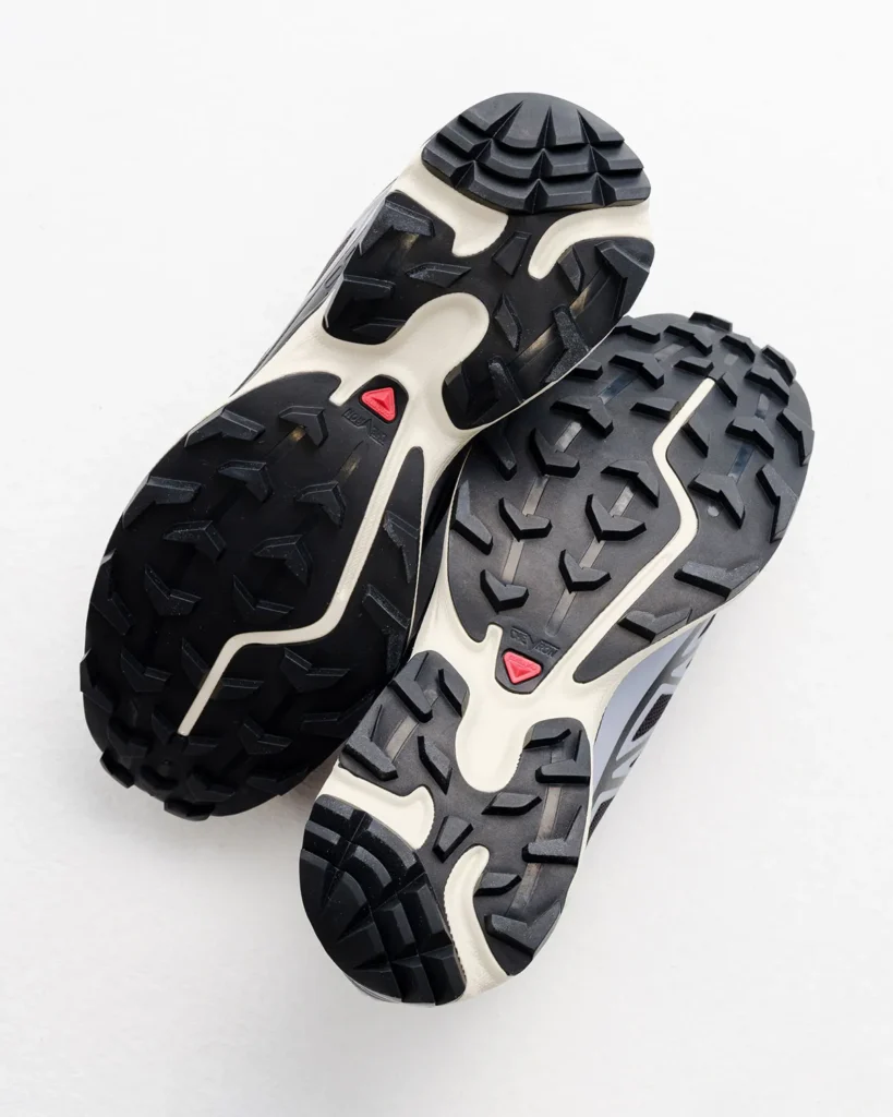 Two trail shoe outsoles overlap on a white background, showing black lugged treads, cream midsoles and red logos
