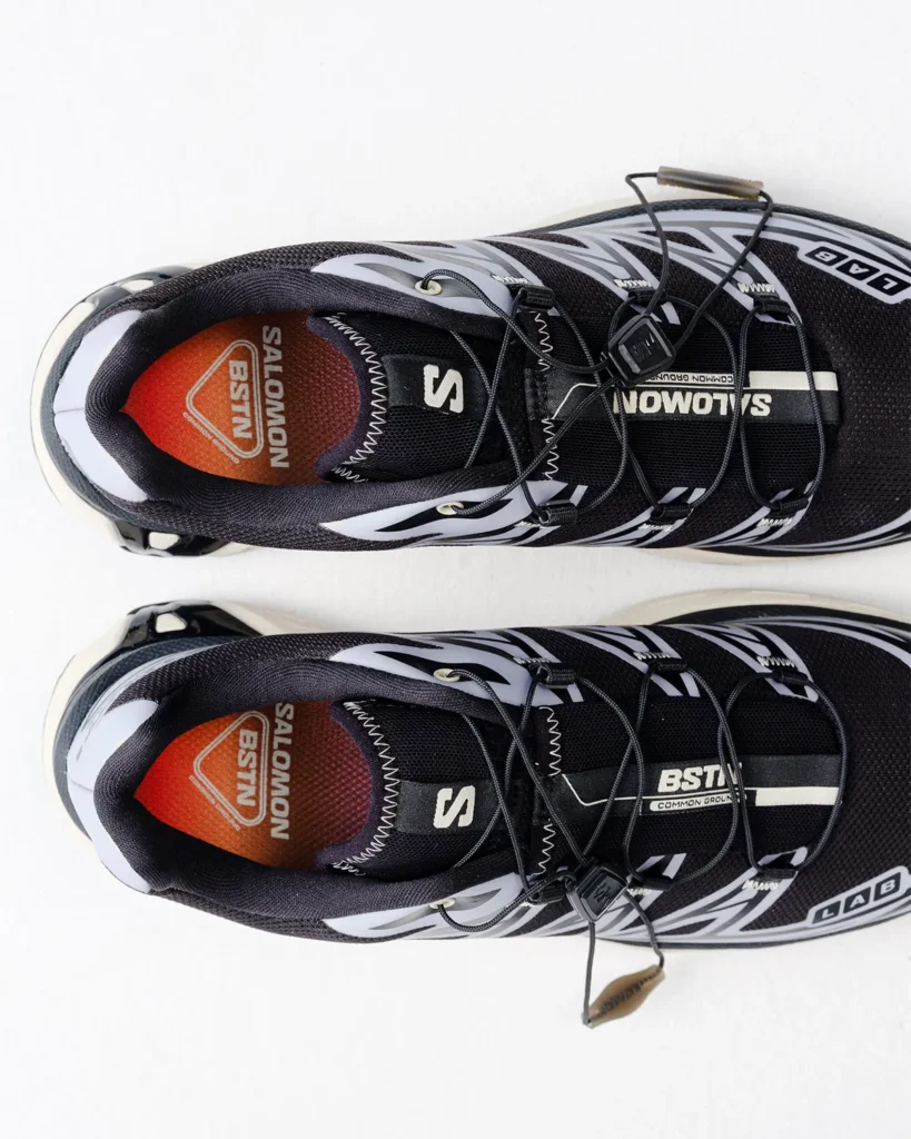 Top-down view of black-and-white Salomon trail shoes with Quicklace systems and orange BSTN-branded insoles on white