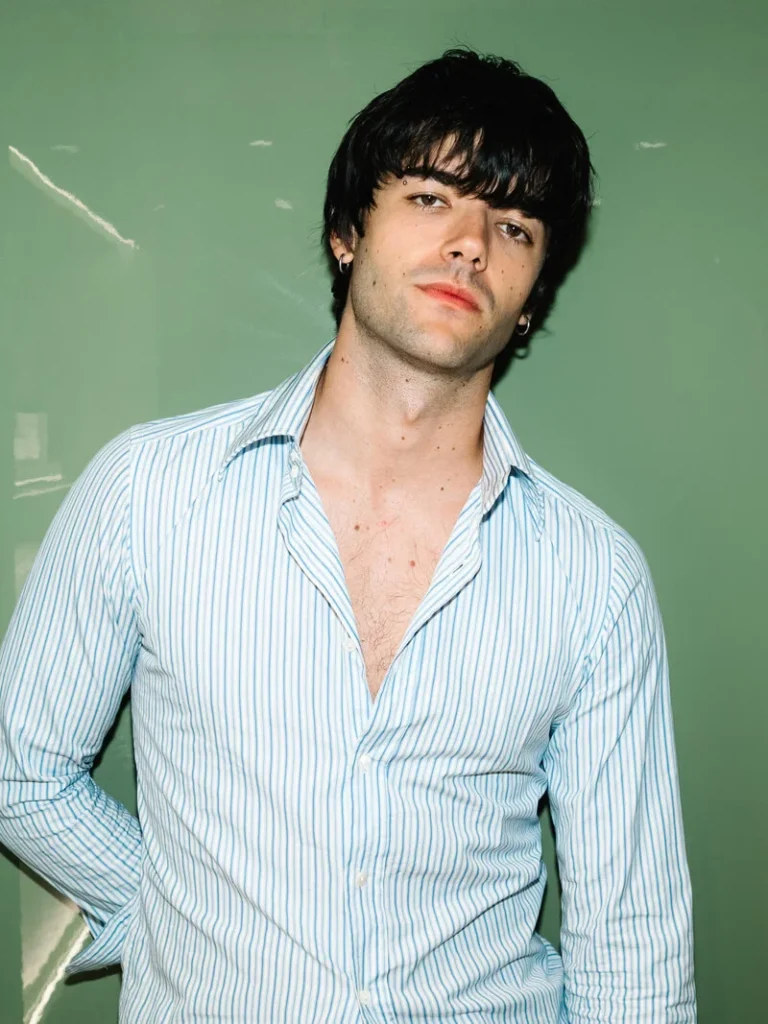 Young man with shaggy dark hair wearing an open blue-and-white striped shirt against a pale green wall