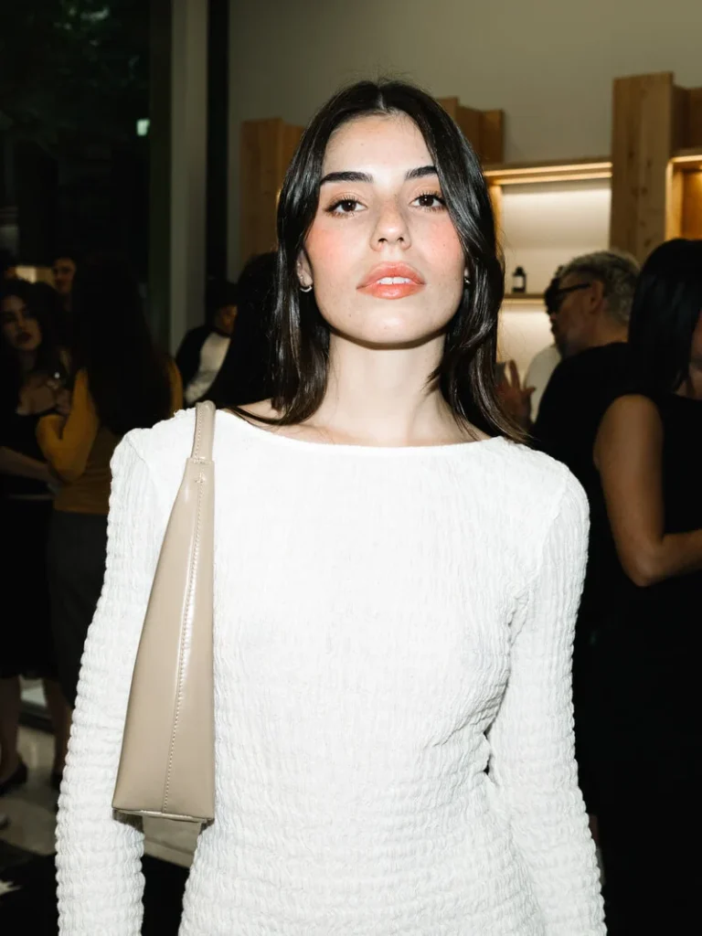 Woman with dark hair wearing a white textured top and carrying a beige shoulder bag at an indoor event
