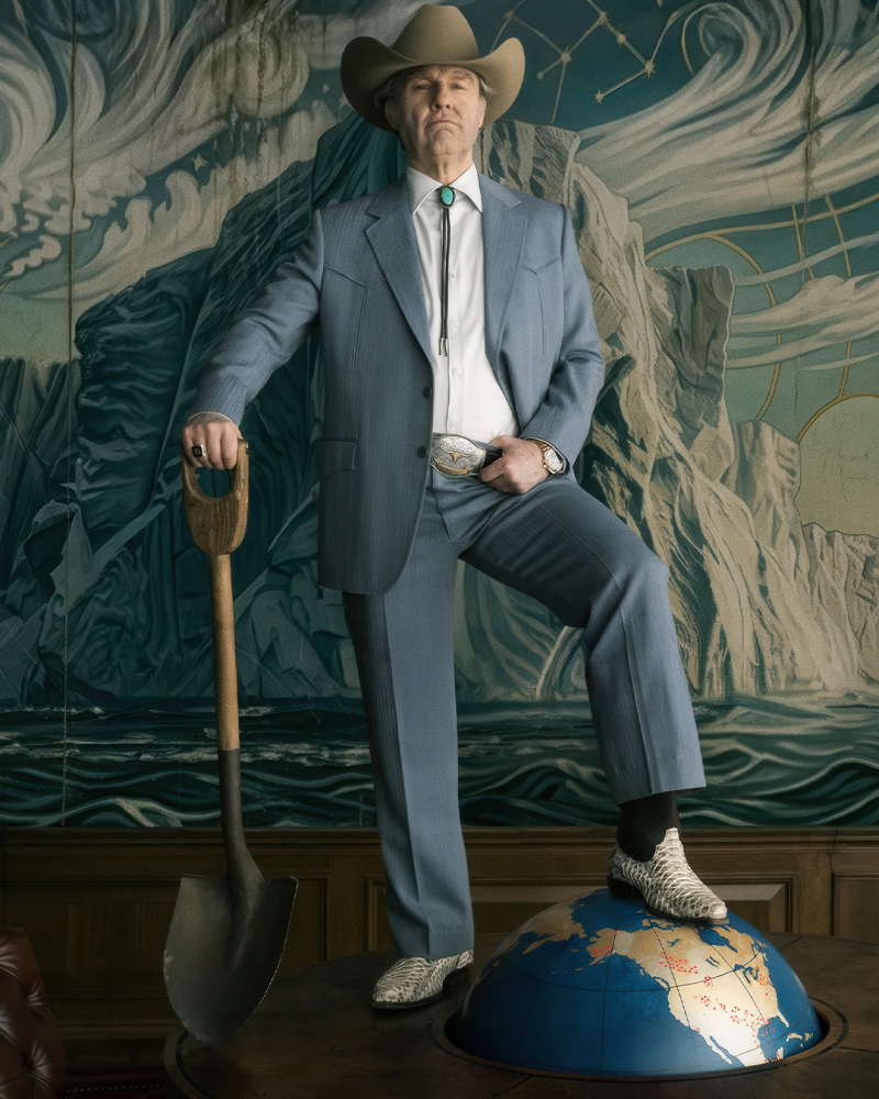 Man in a cowboy hat and blue suit stands with one foot on a globe, holding a shovel before a mountain mural