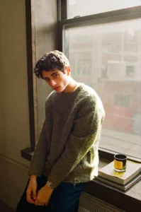 Dom Innarella leans beside a sunlit window in a fuzzy olive-green sweater, with stacked books and a candle nearby