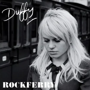 Duffy with long blonde hair looks down, wearing a dark collared jacket, in a black-and-white album cover portrait