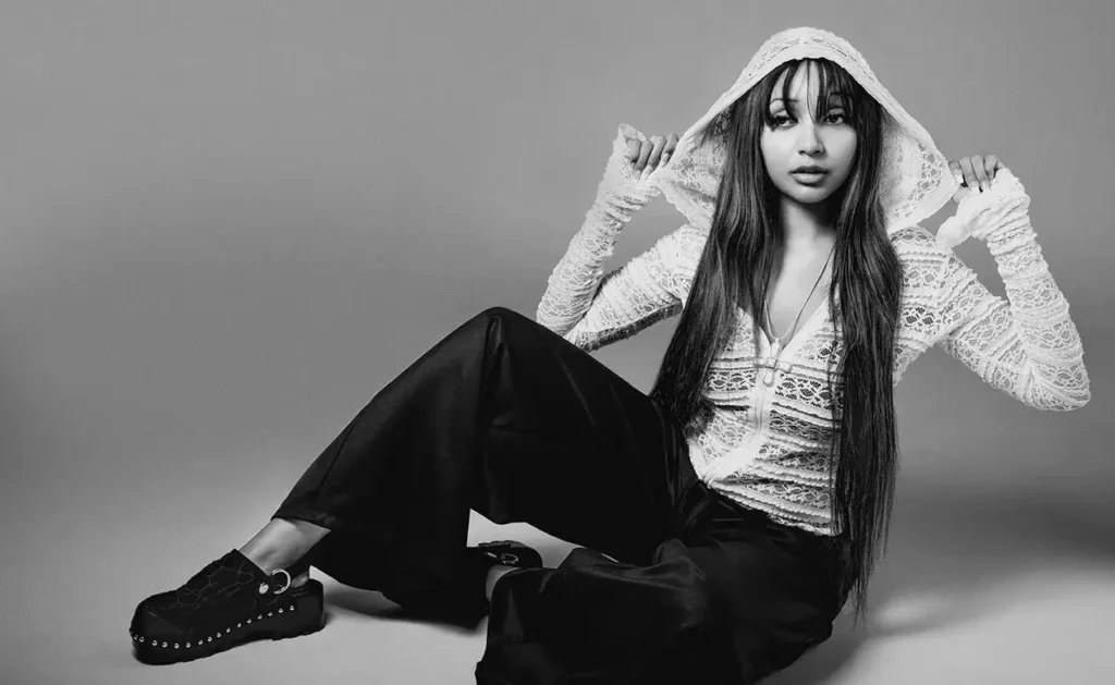 PinkPantheress sits on the floor in a lace hooded top, wide black pants and studded platform shoes