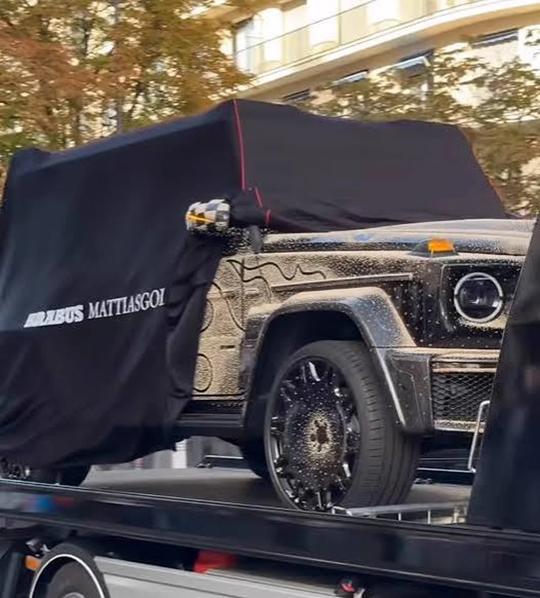 Partially covered black Brabus G-Class with a textured metallic finish on a trailer in an urban setting