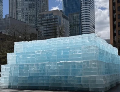 Large stepped structure built from translucent blue ice blocks, with glass skyscrapers and leafless trees behind it