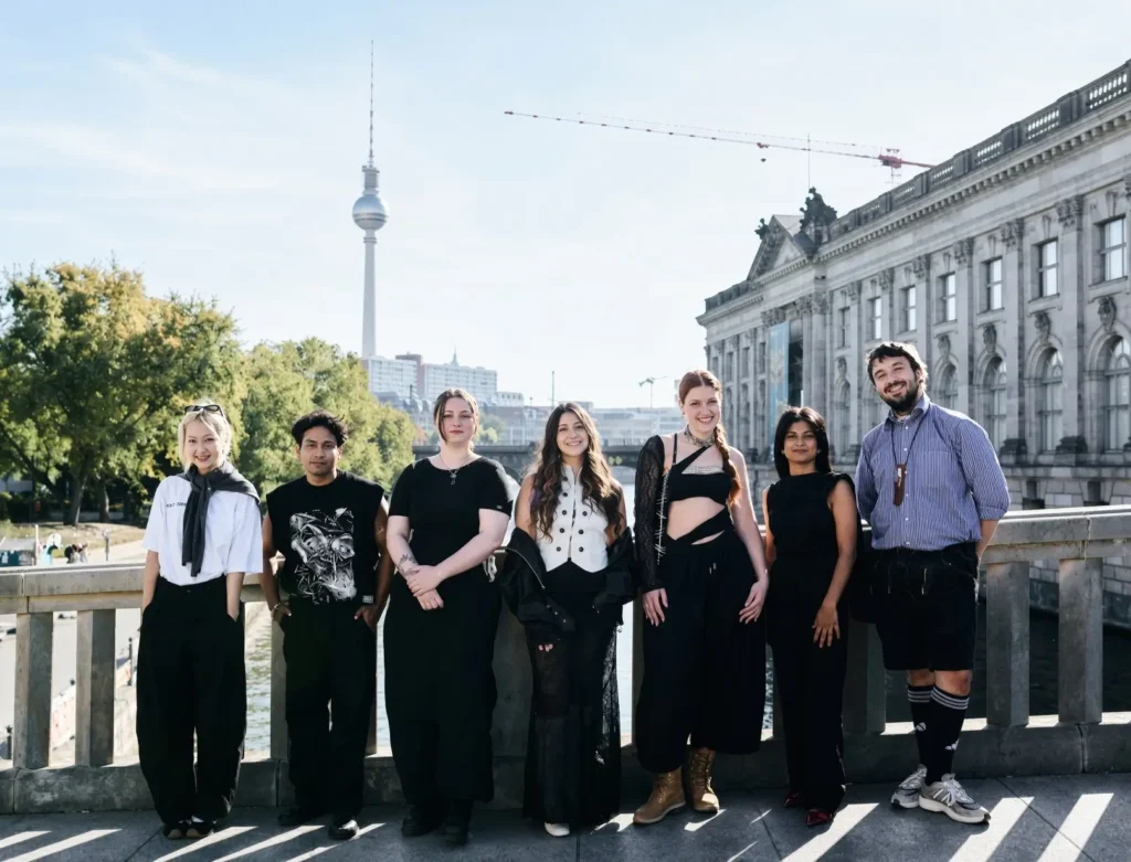 Seven people stand on a bridge in Berlin, wearing mostly black outfits, with the Fernsehturm and ornate building behind them