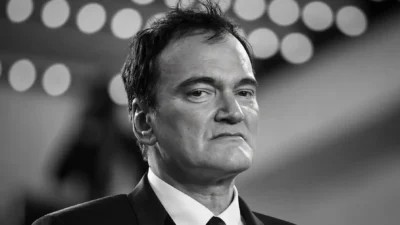 Close-up black-and-white portrait of Quentin Tarantino in a suit, with blurred circular theater lights behind him