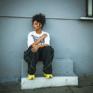 Jessie Reyez sits on concrete steps in a white graphic T-shirt, black pants, and bright yellow shoes