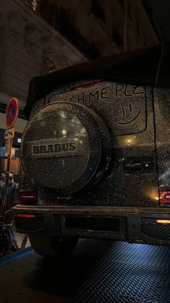 Rain-speckled black BRABUS G-Class rear with graffiti, spare tire cover, and Paris street signs at night