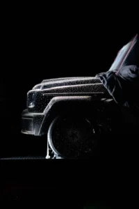 Close-up of a black Mercedes G-Class front with raindrops, partly obscured by a person in a dark jacket against black