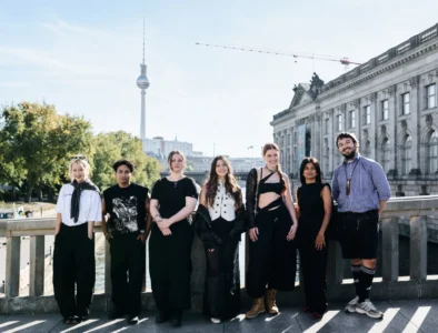 Seven people stand on a bridge in Berlin, wearing mostly black outfits, with the Fernsehturm and ornate building behind them