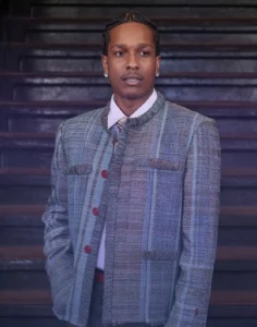 A$AP Rocky stands in front of dark wood paneling, wearing a gray textured jacket, white shirt, tie, and diamond earrings