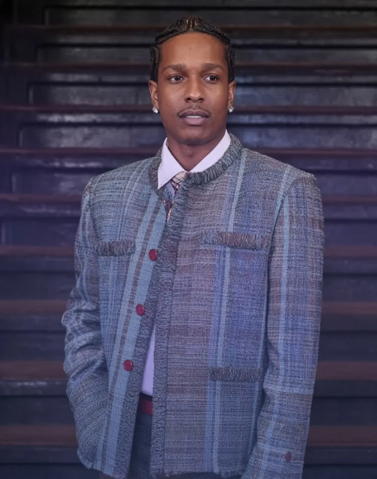 A$AP Rocky stands in front of dark wood paneling, wearing a gray textured jacket, white shirt, tie, and diamond earrings