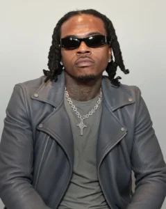 Gunna sits facing the camera in black sunglasses, a gray leather jacket, gray shirt, and layered diamond chains