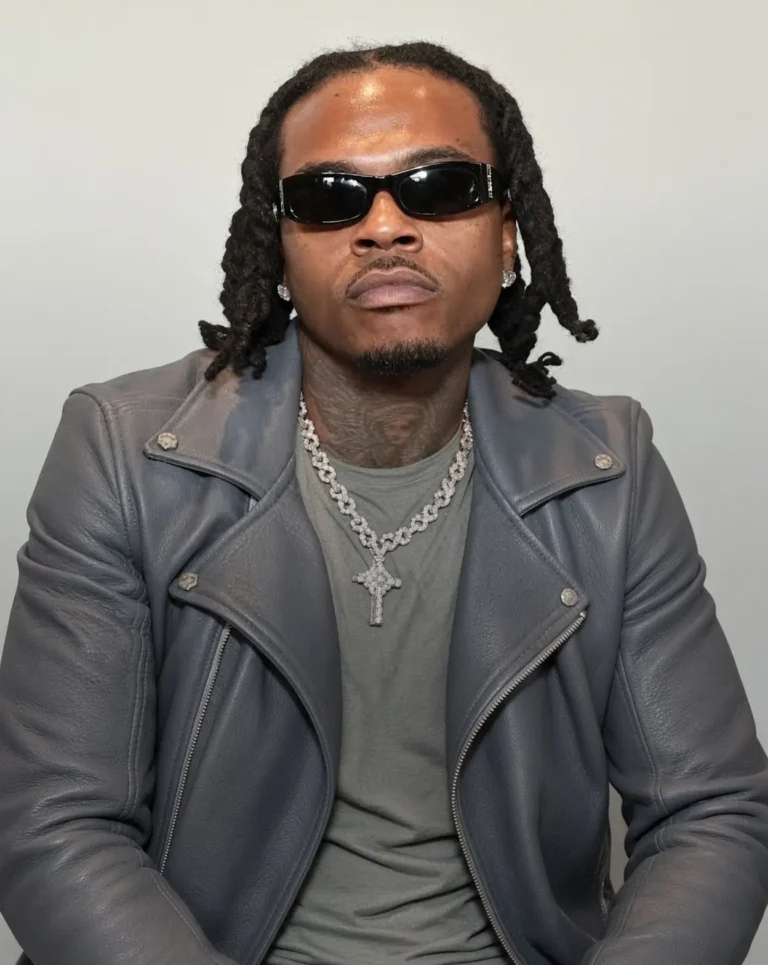 Gunna sits facing the camera in black sunglasses, a gray leather jacket, gray shirt, and layered diamond chains