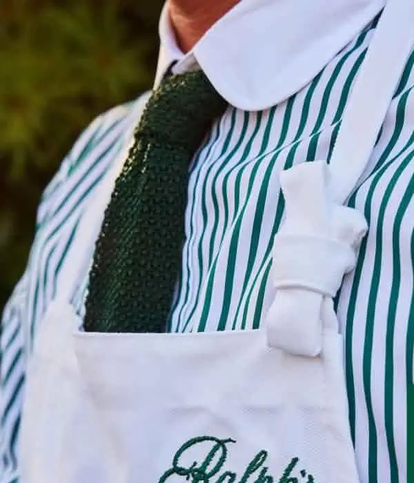 Close-up of a person in a green-and-white striped shirt, textured green tie, white bow, and apron with embroidered logo