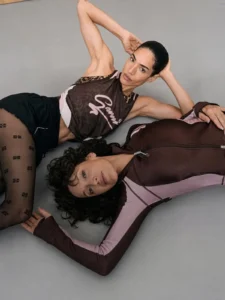 Two models pose on a gray studio floor in brown and pink athletic wear, one lying down and one with arms raised overhead