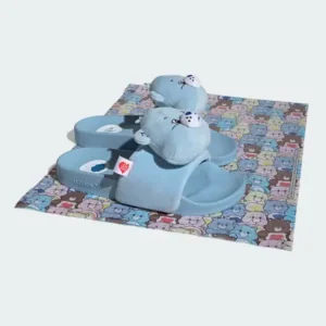 Blue plush animal slippers with white faces on a colorful cartoon-patterned mat against a pale background