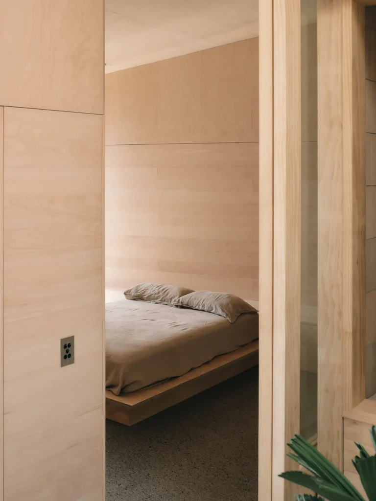 Bedroom with a low wooden platform bed, beige bedding, pale wood walls and a polished concrete floor viewed through a doorway