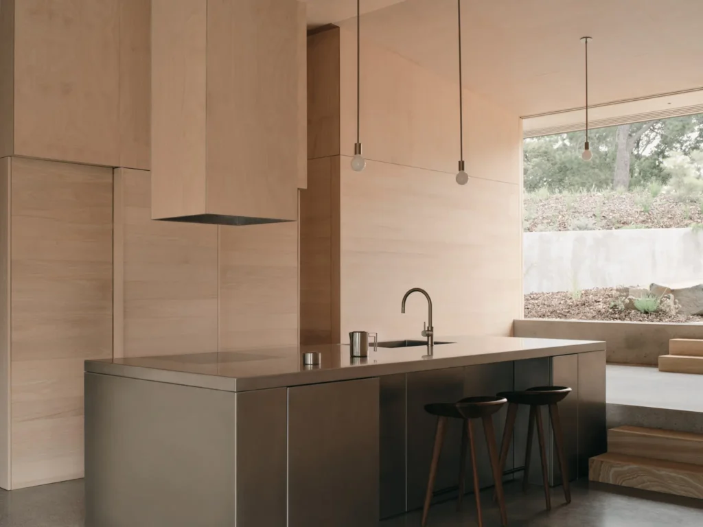 Wood-paneled kitchen with a large island, stainless-steel countertop, black stools, pendant lights and garden views through