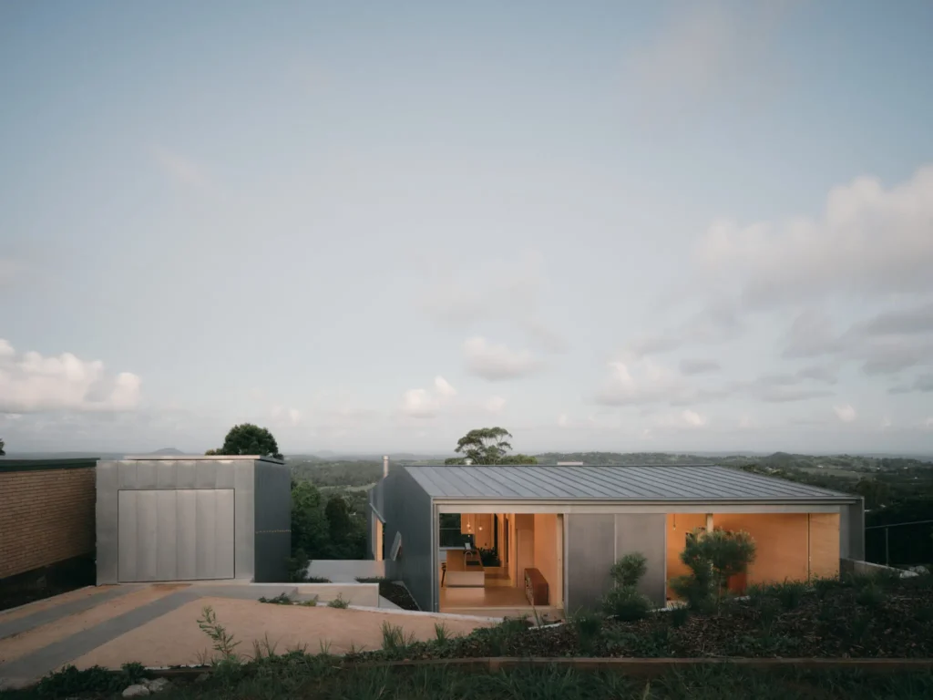 Two zinc-clad pavilions with pitched roofs sit amid native landscaping, overlooking a green Queensland valley under a cloudy